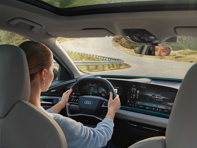 Innensicht eines Audi Q6 e-tron mit Displays, Lenkrad und Fahrerin, die eine kurvige Straße fährt.