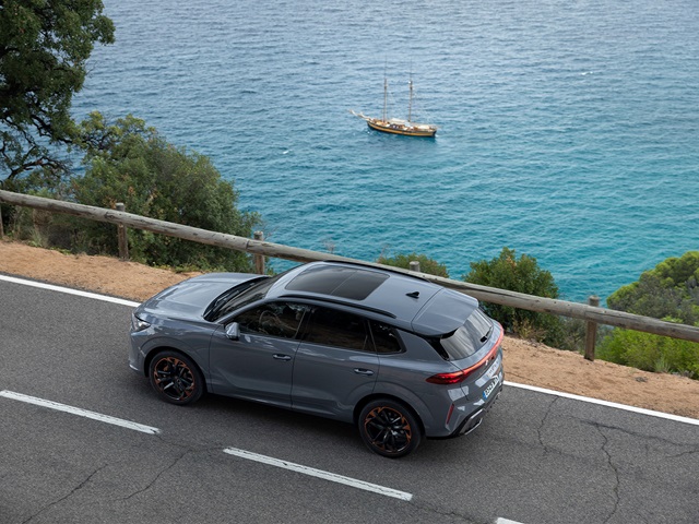 Seitliche Ansicht eines grauen CUPRA Terramar SUV auf einer Küstenstraße mit Meer und Segelboot im Hintergrund