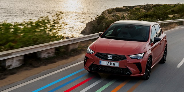 Roter SEAT Ibiza fährt auf Küstenstraße mit Wasser und Felsen im Hintergrund, Frontansicht mit schwarzem Kühlergrill und Nummernschild 'HAHN'