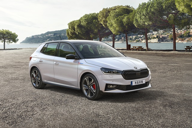 Weiße Škoda Fabia mit schwarzem Kühlergrill und roten Bremssätteln steht seitlich auf einem Parkplatz vor einer Küstenlandschaft mit Bäumen und Bergen