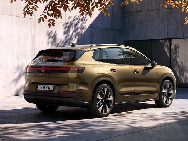Bronzefarbener VW ID. Tiguan von hinten rechts vor einer Betonwand mit Schatten und herabhängendem Laub