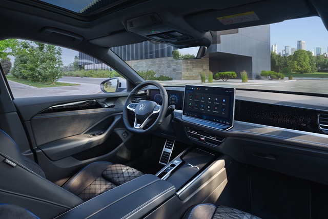 Modern gestalteter Innenraum eines Autos mit großem Touchscreen, digitalem Cockpit und Ledersitzen in Rautensteppung, Blick durch die offene Fahrertür auf Gebäude und Grünfläche.