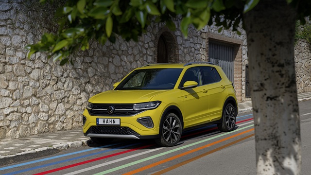 VW T-Cross R-Line in Gelb aus schräger Frontansicht auf einer Straße mit bunten Streifen, im Hintergrund Steinmauer und Tor, im Vordergrund steht ein Baum.