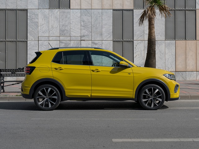 Gelber VW T-Cross R-Line in der Seitenansicht auf einer Straße, im Hintergrund ein modernes Gebäude und eine Palme.
