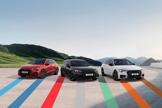 Audi Gebrauchtwagen :plus Wochen – Drei sportliche Audi-Fahrzeuge mit den bunten, dynamischen Linien des Hahn Codes auf einem Parkplatz vor einer Bergkulisse.