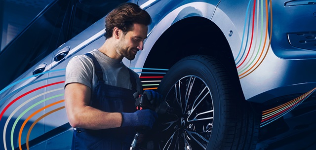 Mann in blauer Arbeitskleidung montiert mit einem Werkzeug einen Reifen an einem Auto.